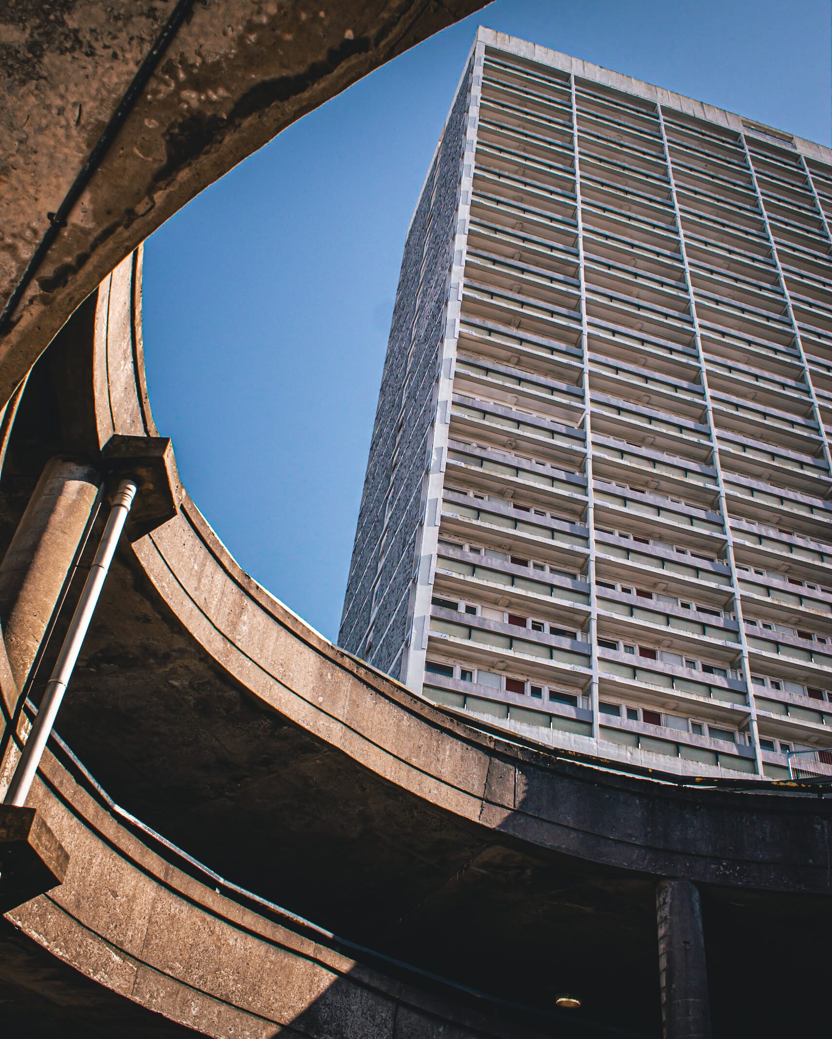 A tall, grey high-rise tower framed by the curved concrete ramp of a multi-storey car park, set against a clear blue sky. The view captures strong architectural lines and contrasting shapes.