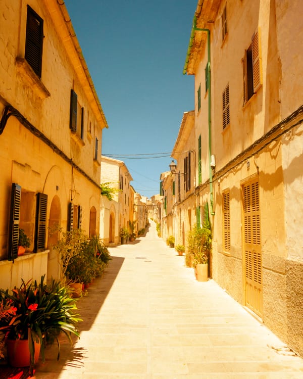 Empty sunlit street with shutters and potted plants in Alcúdia Old Town, Mallorca.
