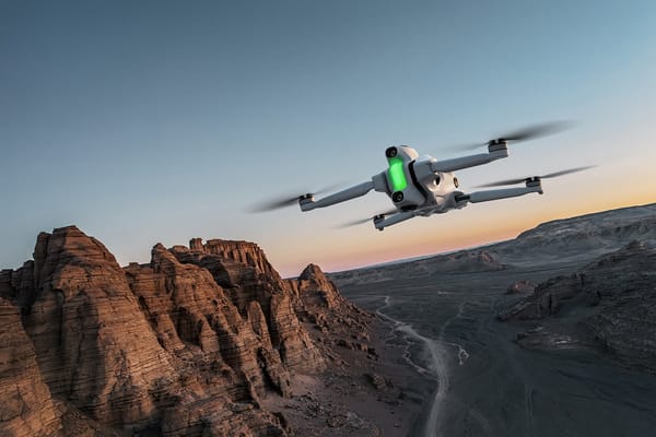 White Antigravity A1 drone with a green light flies over rocky desert cliffs at sunset, wide sky and empty valley.